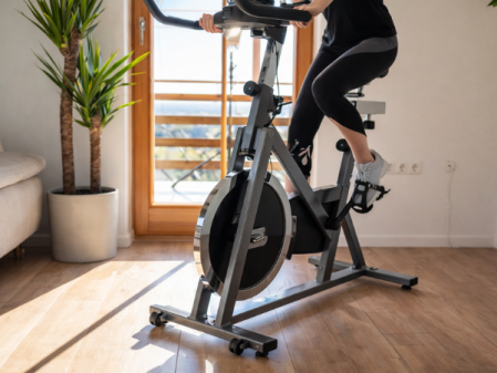 Persona usando una bicicleta estática en un salón luminoso, con suelo de madera, luz natural entrando por una puerta acristalada y plantas decorativas alrededor